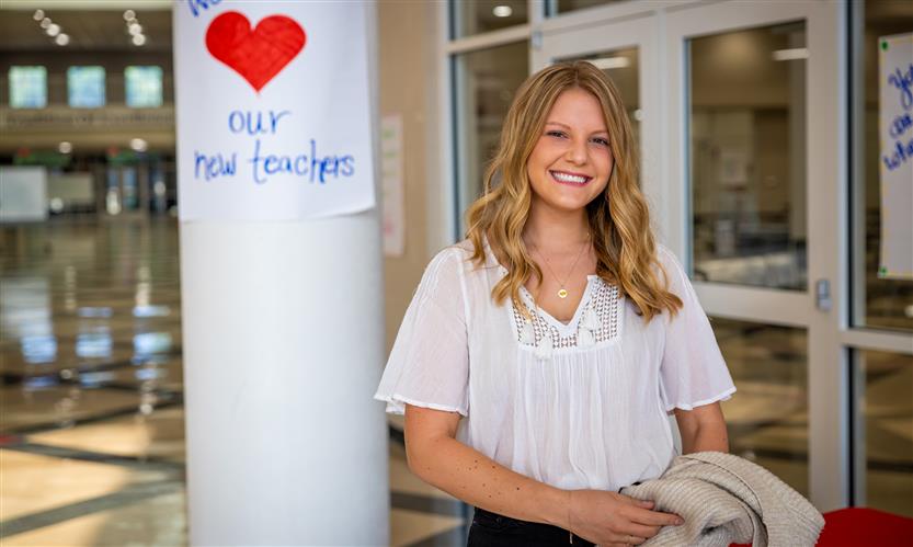 A woman wearing a white shirt smiles while standing on front of a sign that reads \